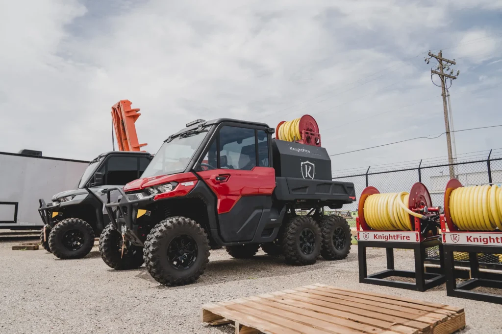 Two off-road fire protection vehicles with mounted yellow hose reels parked near a chain-link fence.