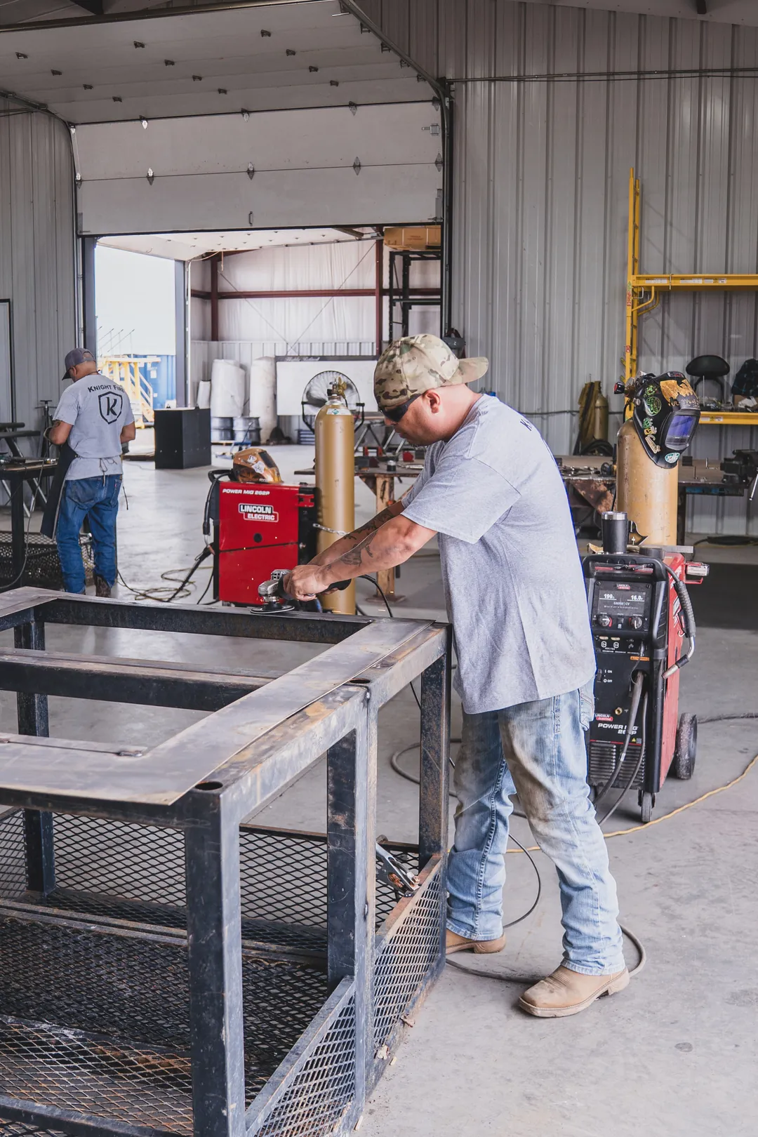 Man wearing a cap and safety glasses uses a grinder on a large metal frame inside a workshop.