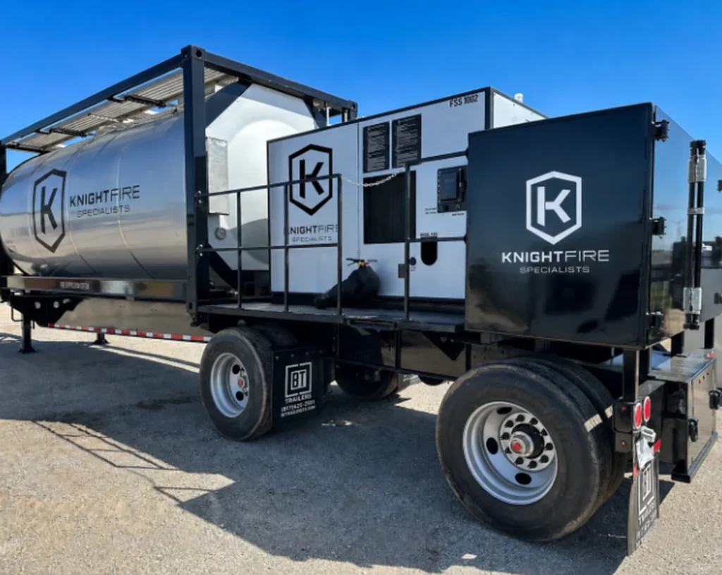 A large trailer with silver and black compartments labeled KnightFire Specialists parked on gravel under a clear blue sky.
